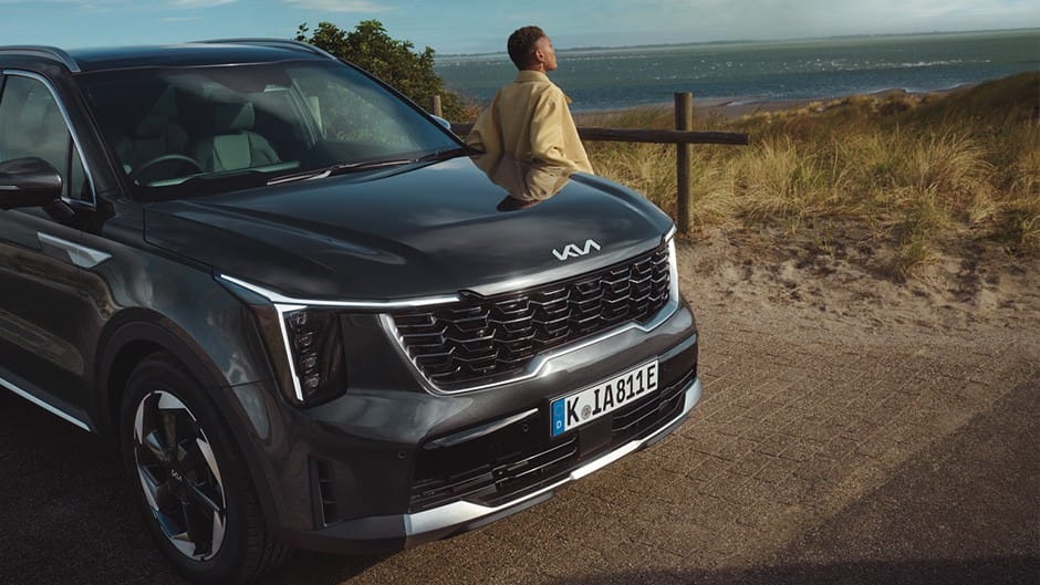 The Kia Sortento ‘4’ in Midnight Black parked on a road overlooking a green landscape, with a lady leaning against it.