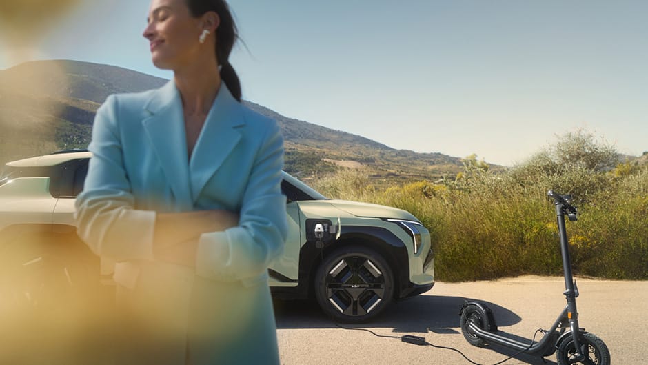 A Kia GT-Line S EV3 in Pistachio Green, with an electric scooter which has been plugged into the car. The car is positioned in front of a landscape with a mountain in the background, and a women stood in front of the EV3 with her head turned left.