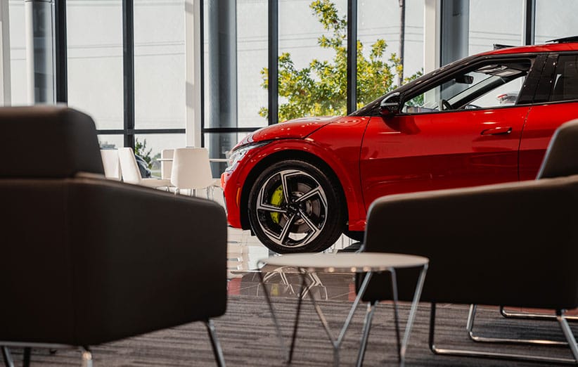 Front section of a red Kia EV6 parked in a car dealer network location lobby, with windows, plants, chairs, and tables visible in the background and foreground.
