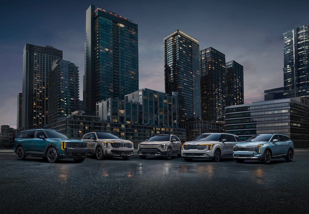 Five modern, impressive Kia vehicles parked on a street, in blue, tan, and white, reflecting Kia's hybrid SUV lineup. The cityscape is visible in the distance, with tall buildings towering over the cars at late sunset.