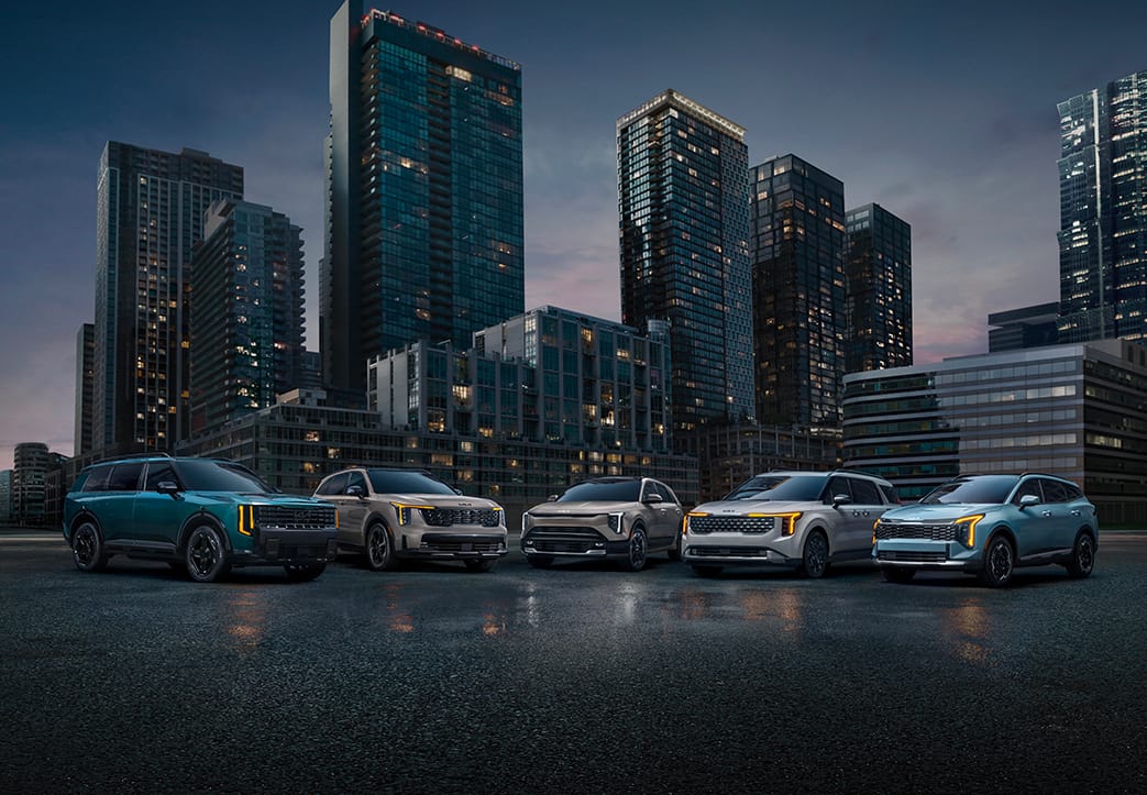 Five modern, impressive Kia vehicles parked on a street, in blue, tan, and white, reflecting Kia's hybrid SUV lineup. The cityscape is visible in the distance, with tall buildings towering over the cars at late sunset.
