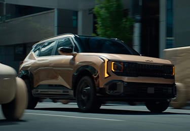 The all-new 2027 Kia Seltos in tan driving with a tan concept model in the lane next to it on an urban street with buildings and trees behind the vehicles, highlighting the SUV’s sleek exterior with LED headlights and a black grille.