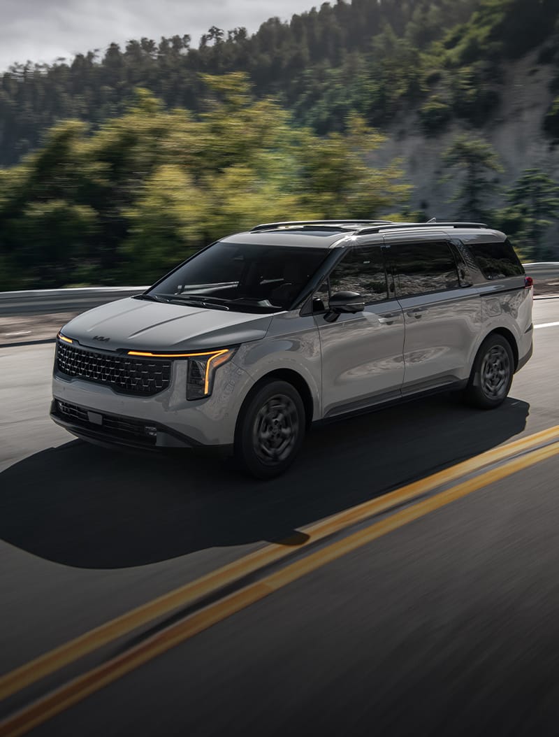 2026 Kia Carnival MPV Hybrid, three-quarter driver's-side view, driving on a road in the mountains.