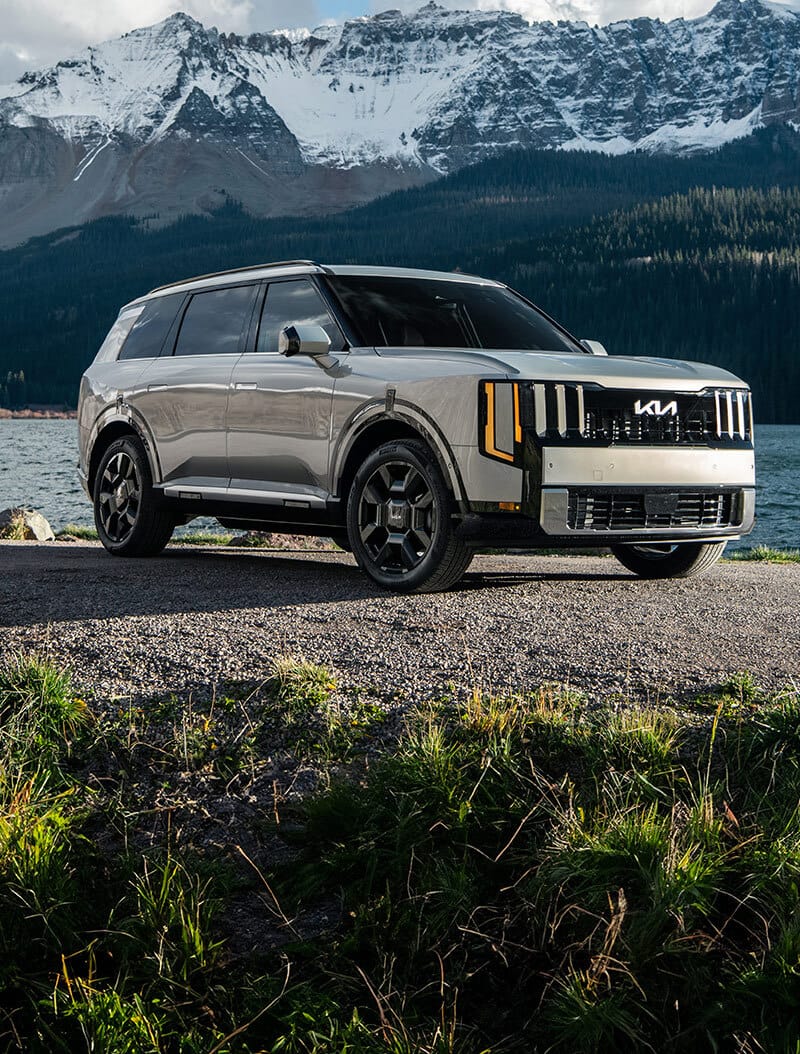 Un Kia Telluride Hybrid 2027 plateado estacionado junto a un lago rocoso con imponentes montañas cubiertas de nieve al fondo, destacando el audaz diseño exterior y las capacidades robustas del SUV.