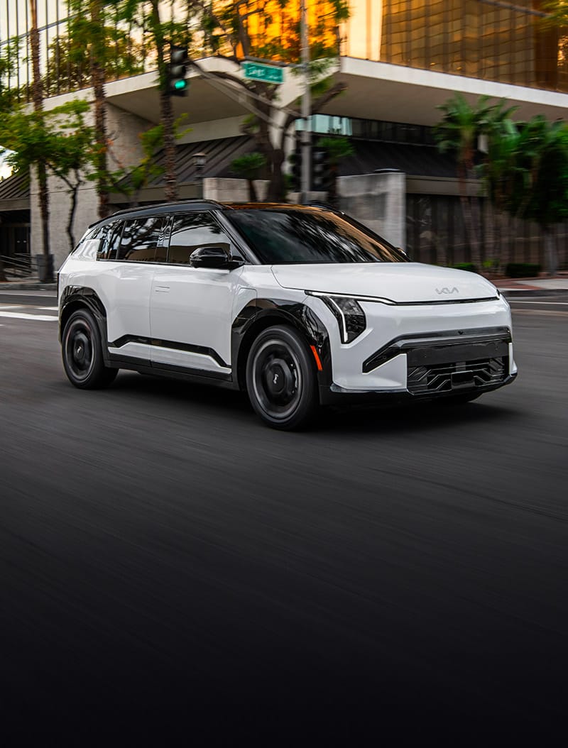 An action shot of the Kia EV3 compact electric SUV in white driving down an urban street, with trees and buildings in the background. The image highlights the sleek design, headlights, and interesting exterior features of the all-new EV.