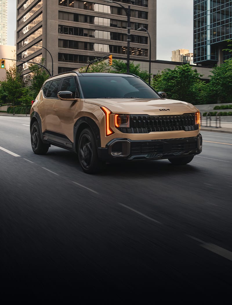 2027 Kia Seltos in brown driving on the street with buildings in the background, shown in a front view with gloss black exterior trim and a bold front grille against an urban landscape.