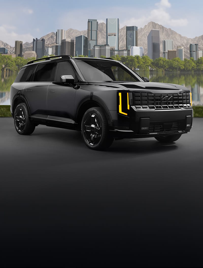 A still image of the 2027 Kia Telluride in black parked on a street with a lake, tall buildings, and mountains behind it, highlighting the front and passenger side of the SUV, the exclusive wheels, and the LED headlights.