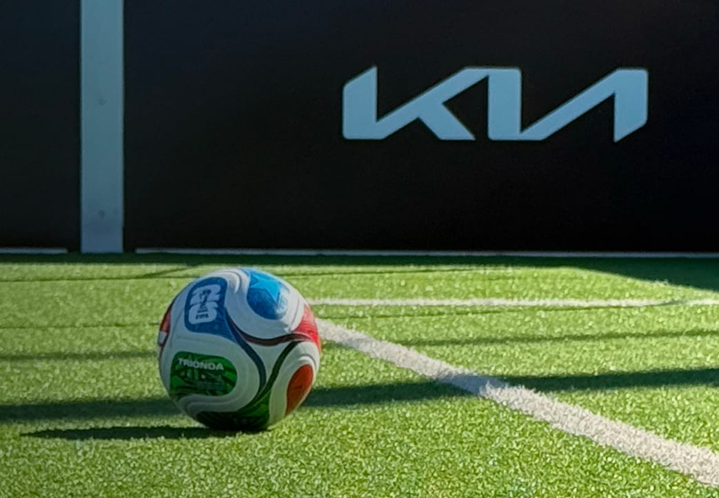 An atmospheric image of a colorful soccer ball on a green, white-lined soccer turf field, with FIFA World Cup 2026 and Kia Official Partner branding visible in the background.