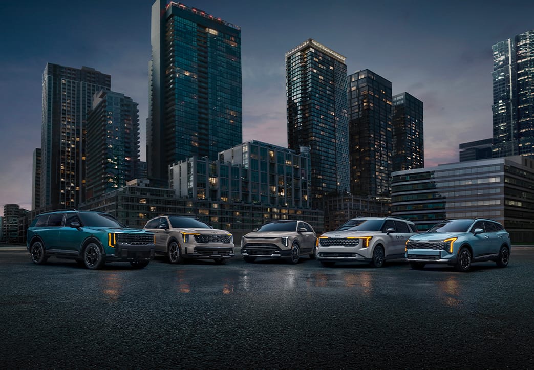 Five modern, impressive Kia vehicles parked on a street, in blue, tan, and white, reflecting Kia's hybrid SUV lineup. The cityscape is visible in the distance, with tall buildings towering over the cars at late sunset.