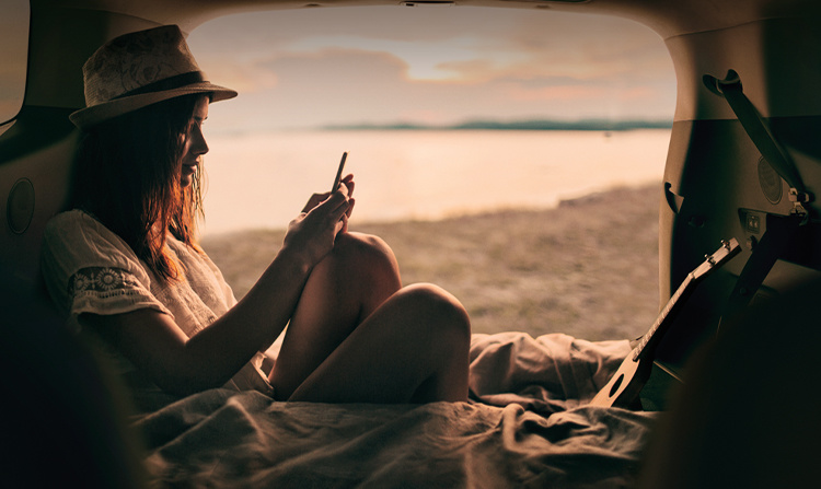 Persona sentada mientras usa su teléfono en un vehículo Kia estacionado en la playa al atardecer