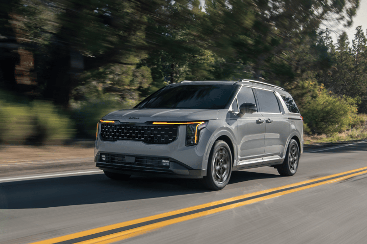 2025 Kia Carnival MPV Hybrid, three-quarter front view, driving on a road in the mountains featuring the signature vertical LED headlights