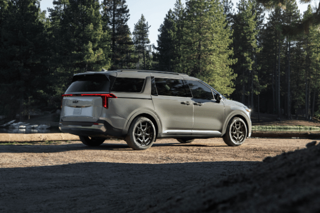 2025 Kia Carnival MPV Hybrid, three-quarter back view, parked by a lake in the forest