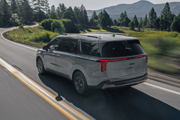 2025 Kia Carnival MPV Hybrid, three-quarter back view, driving on a road by lush trees featuring vertical LED tail lights