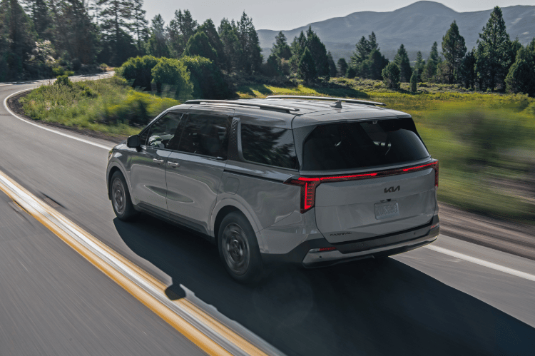 2025 Kia Carnival MPV Hybrid, three-quarter back view, driving on a road by lush trees featuring vertical LED tail lights