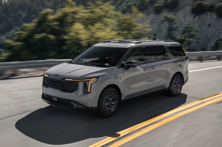 2025 Kia Carnival MPV Hybrid, three-quarter driver's side view, driving on a road in the mountains featuring the signature vertical LED headlights
