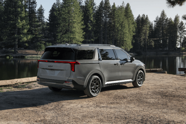 2025 Kia Carnival MPV Hybrid, three-quarter back view, parked by a lake in the forest