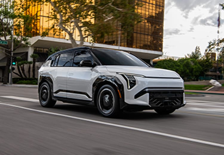 Photograph of the 2027 Kia EV3 compact electric SUV in grey-white driving down an urban street, with trees and buildings in the background. The image highlights the sleek silhouette and interesting exterior features of the all-new EV.