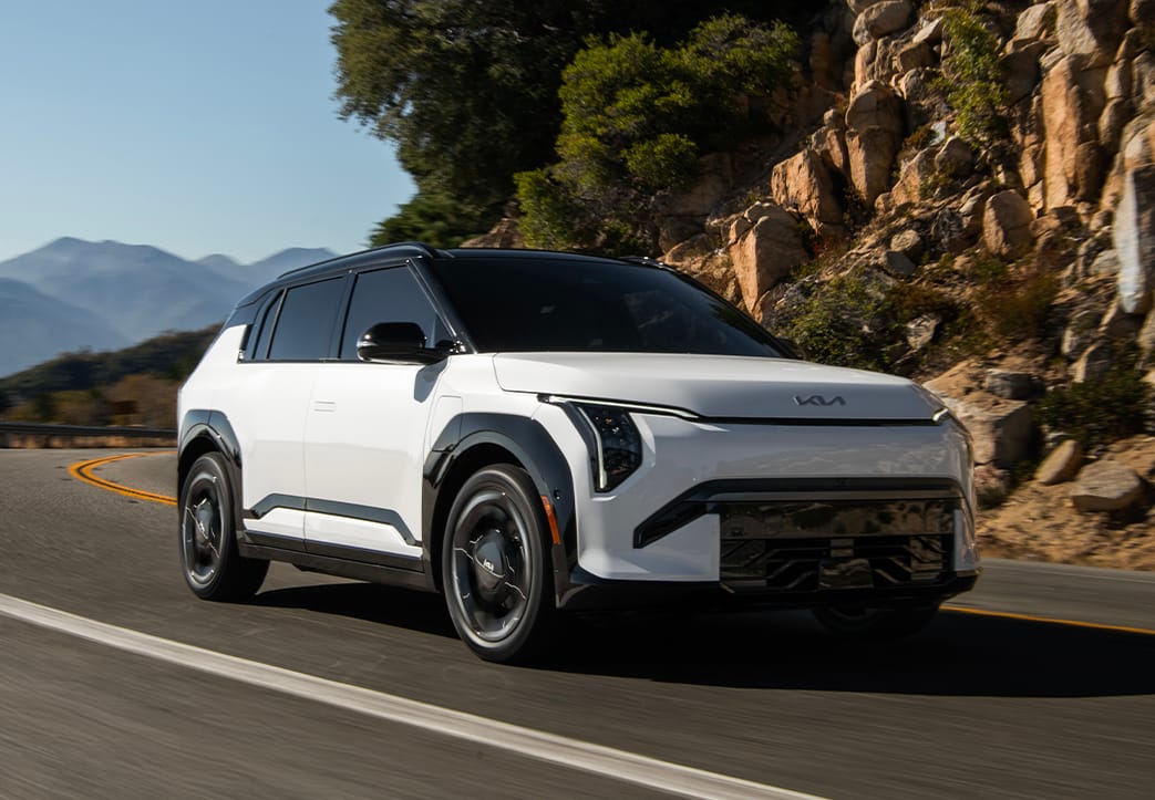 Front angle view of the 2027 Kia EV3 compact electric SUV as it turns a corner on a curvy road with rocky mountains and trees in the background. The front grille and Kia logo are visible as well as the black highlighting on white.
