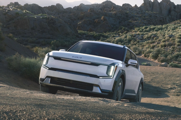 2024 Kia EV9 in white, three-quarter front view, driving up an unpaved road in the dessert