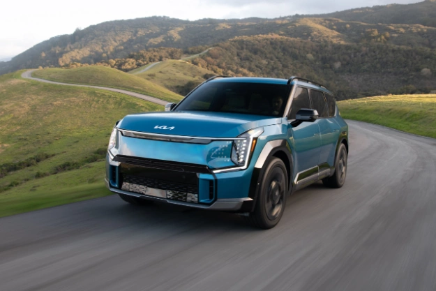 2024 Kia EV9 in blue, three-quarter front view, driving through a paved road in the hills
