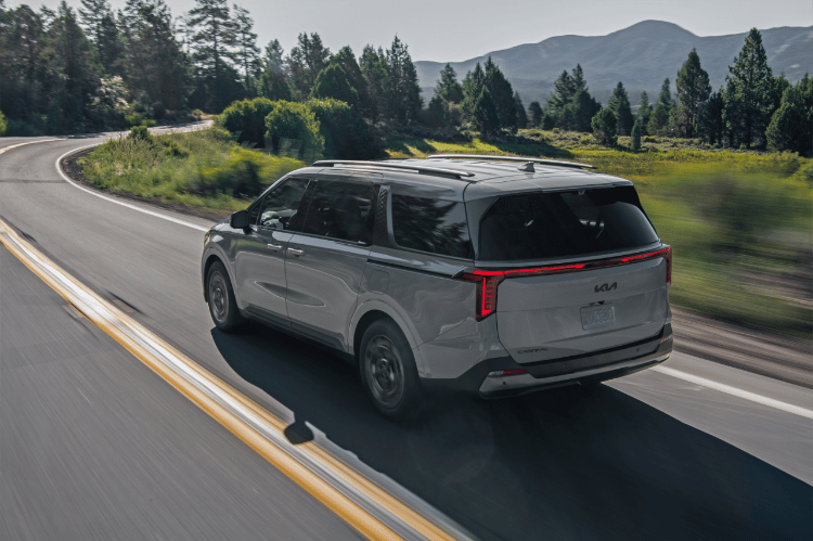 2025 Kia Carnival in gray, driving fast down a curving road near the forest