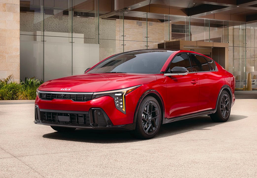 A still shot of a red 2027 Kia K4 in a three-quarter front driver's side view, featuring the sleek front grille. The sedan is parked in a driveway with a modern house with tall windows in the background.