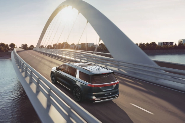 2024 Kia Carnival Driving On A Bridge That Hovers Above A Lake During Sunrise Rear Three-Quarter View 