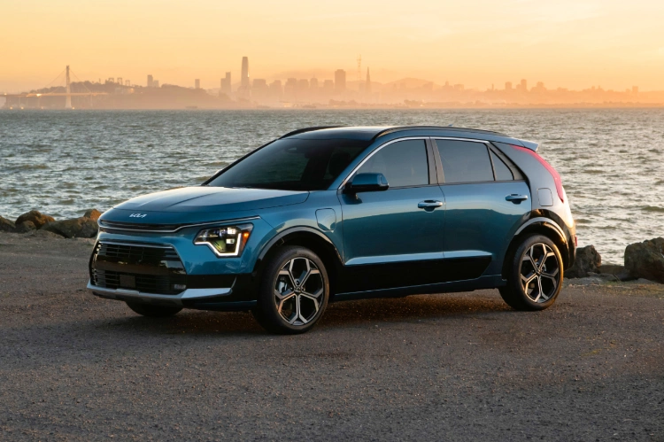 2023 Niro PHEV in blue, parked in front of the ocean at sunset with a city skyline in the distance.
