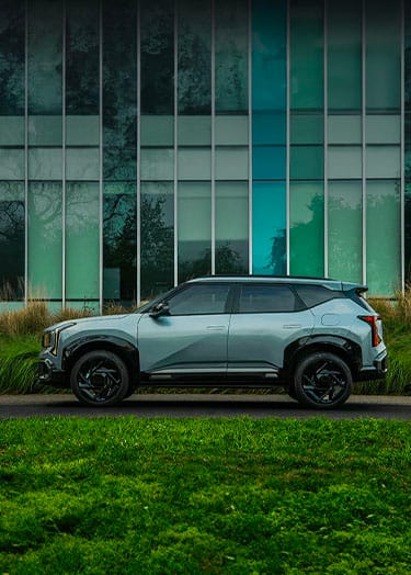 An Iceberg Green 2027 Kia Seltos is parked beside a grassy area, featuring black trim and black alloy wheels. A modern building with tall vertical glass panels and greenery behind them forms the backdrop.
