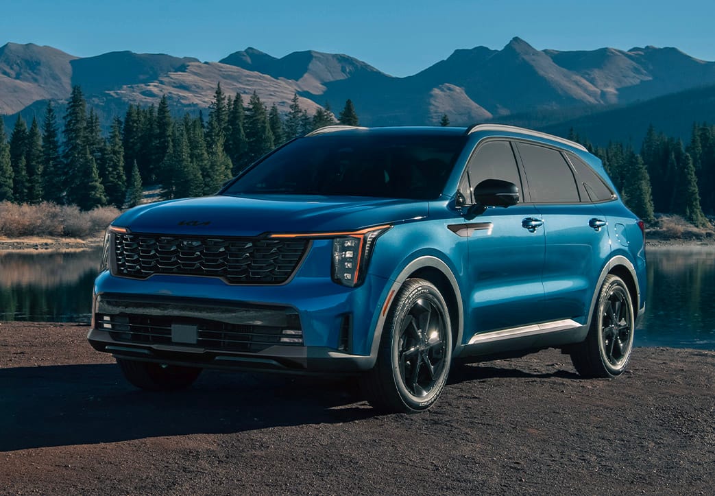 Front driver-side photograph of the 2027 Kia Sorento Hybrid SUV, parked outdoors near a beautiful pond reflecting trees and mountains in the background. The Sorento is shining in the sun, with its sleek profile and finely-designed lines conveying power and elegance.
