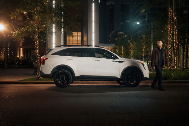 A white 2025 Kia Sorento is parked on a street at night, with a person walking past it