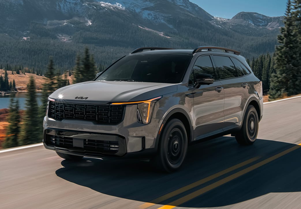 A gray 2027 Kia Sorento in a three-quarter driver's side view, highlighting the LED headlights and front grille. The SUV is driving on a road with a lake, mountains, and greenery next to it.