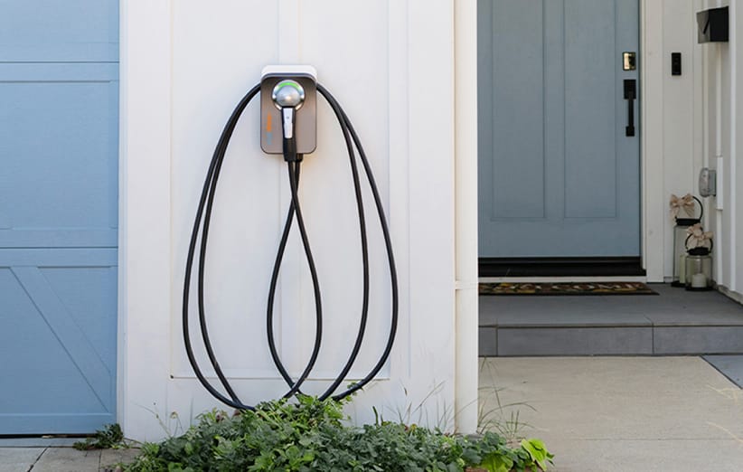 A Level 2 ChargePoint Home Flex hybrid-electric vehicle charging cable and connector, coiled up and attached to the side of a Kia owner's home garage, with shrubs and blue front door in background.