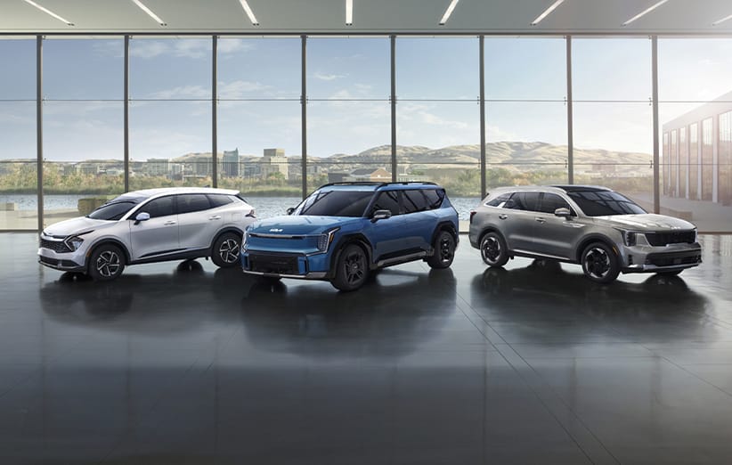 Three Kia SUVs are displayed inside a modern showroom with floor-to-ceiling windows, featuring white, blue, and silver electric vehicles lined up side by side. There are buildings and mountains visible through the windows.