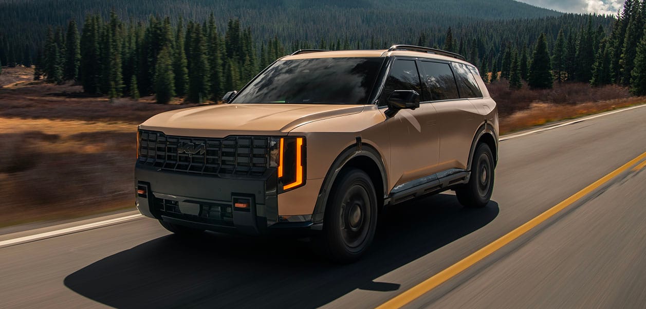Action shot of the 2027 Kia Telluride in brown, highlighting the front and driver-side profile with a bold grille, driving along a road framed by tall green trees.