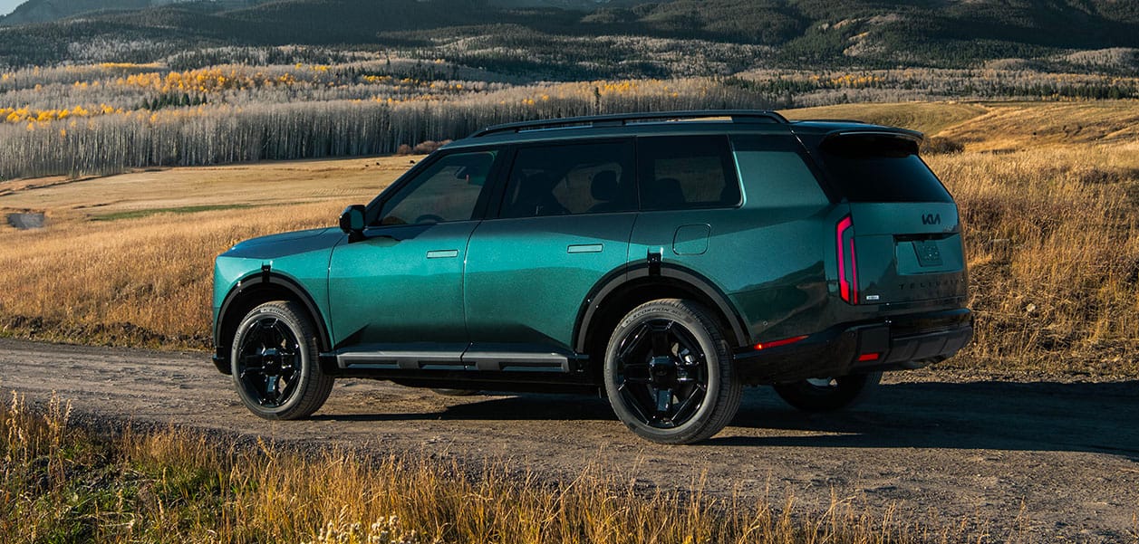 A still shot of the 2027 Hybrid Kia Telluride in blue-green parked on a dirt path, with the driver's side of the SUV facing out against a backdrop of foliage and snowy mountains.