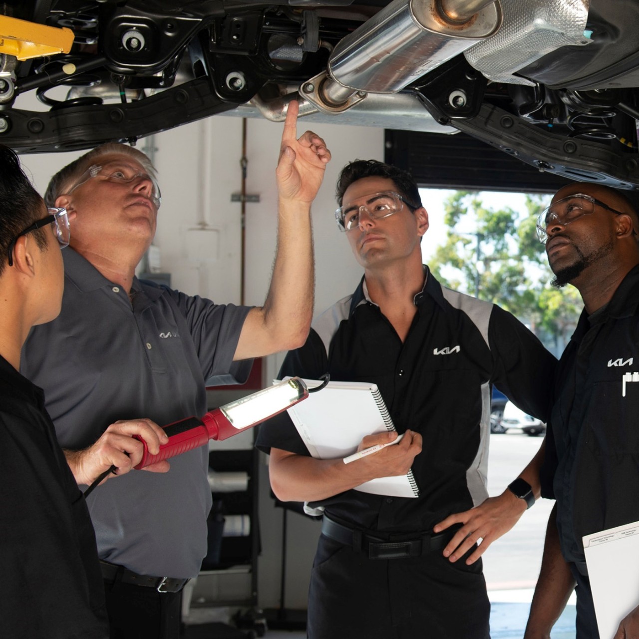 A group of four Kia technicians analyze and discuss a mechanical component from below a raised Kia vehicle.