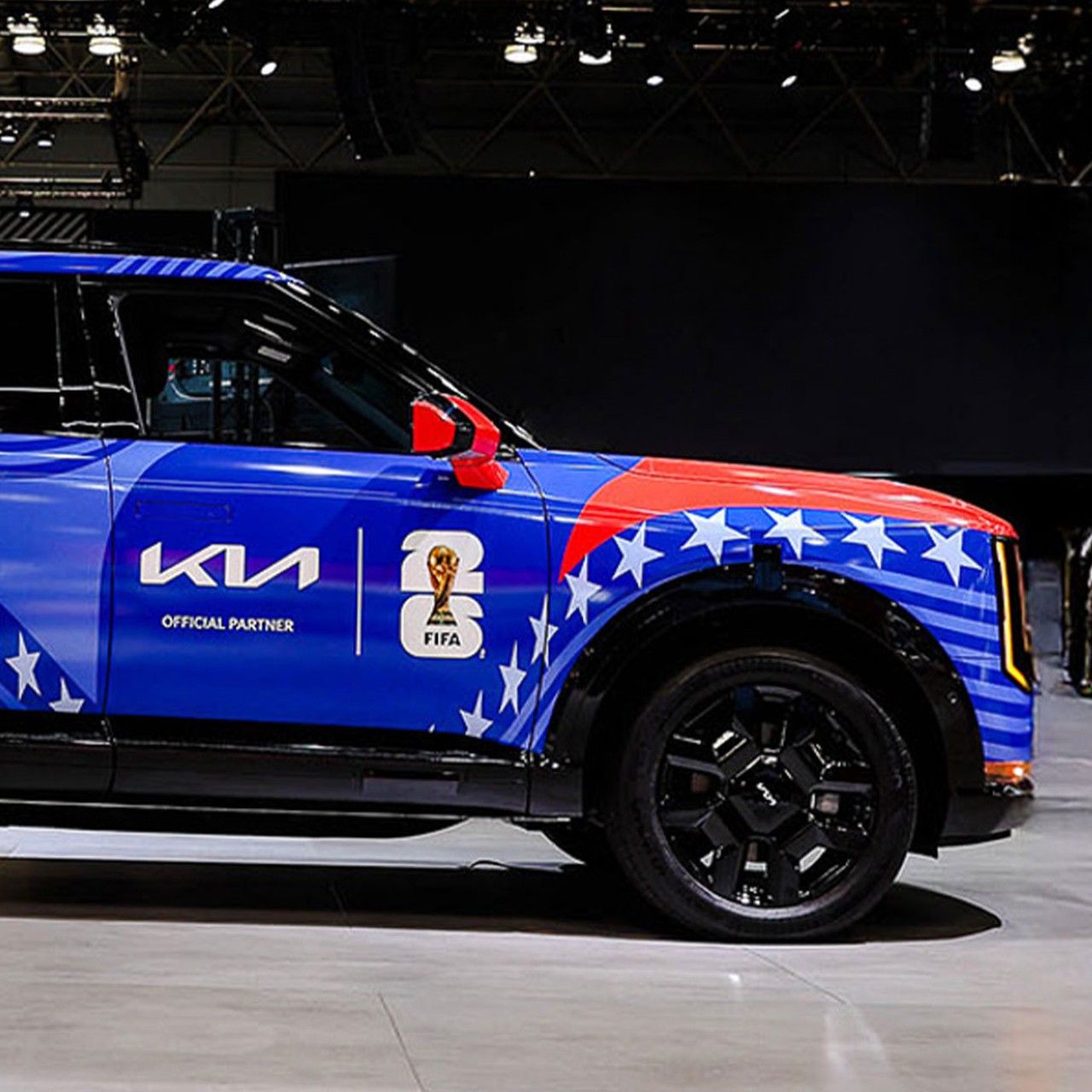 Side profile view of the front section of a Kia SUV, covered in a blue, white, and red wrap with stars, stripes, and FIFA 2026 official branding. The vehicle sits in a brightly-lit showroom.