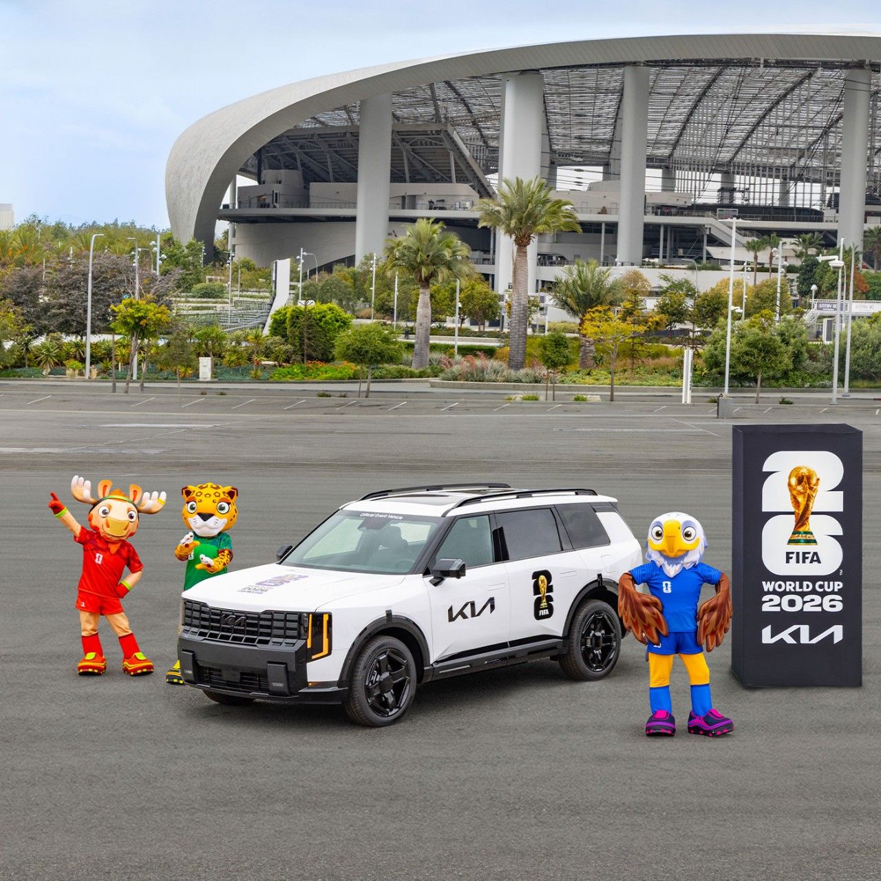A white Kia Telluride HEV parked in front of SoFi Stadium with three FIFA World Cup 2026 mascots and a branded Kia tournament display tower. The stadium's iconic curved canopy roof is visible in the background.