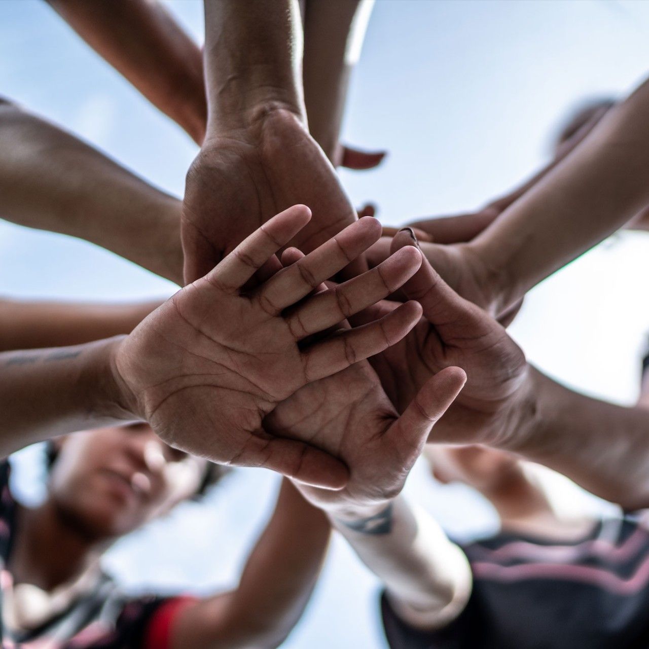 Low angle photograph looking directly upwards at a group of people putting their hands in the ring on top of each others', with a focus on the palms of the hands, meant to convey teamwork and inspiration.