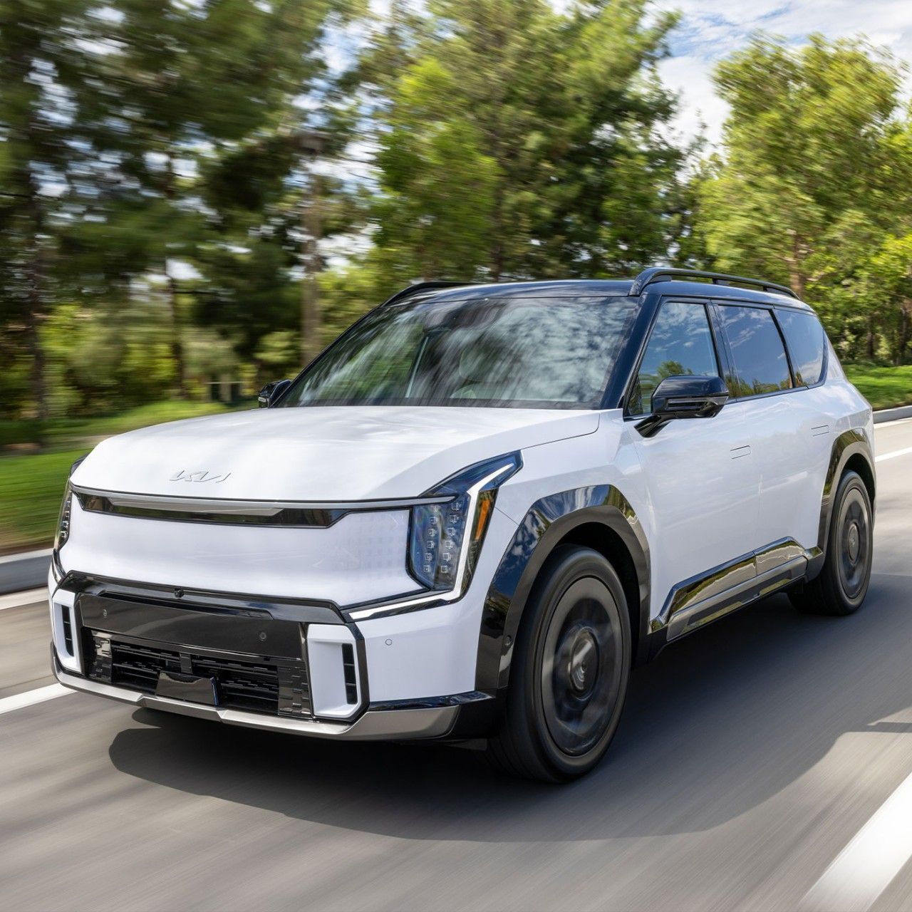 A photograph of a white Kia EV9 electric SUV, highlighting its front grille, headlights, and driver's side angle, as it drives quickly over a paved road with trees and sky in the background blurred from the motion. The photo evokes an official, business-like mood, inspiring the idea that this SUV could be one of multiple company cars.
