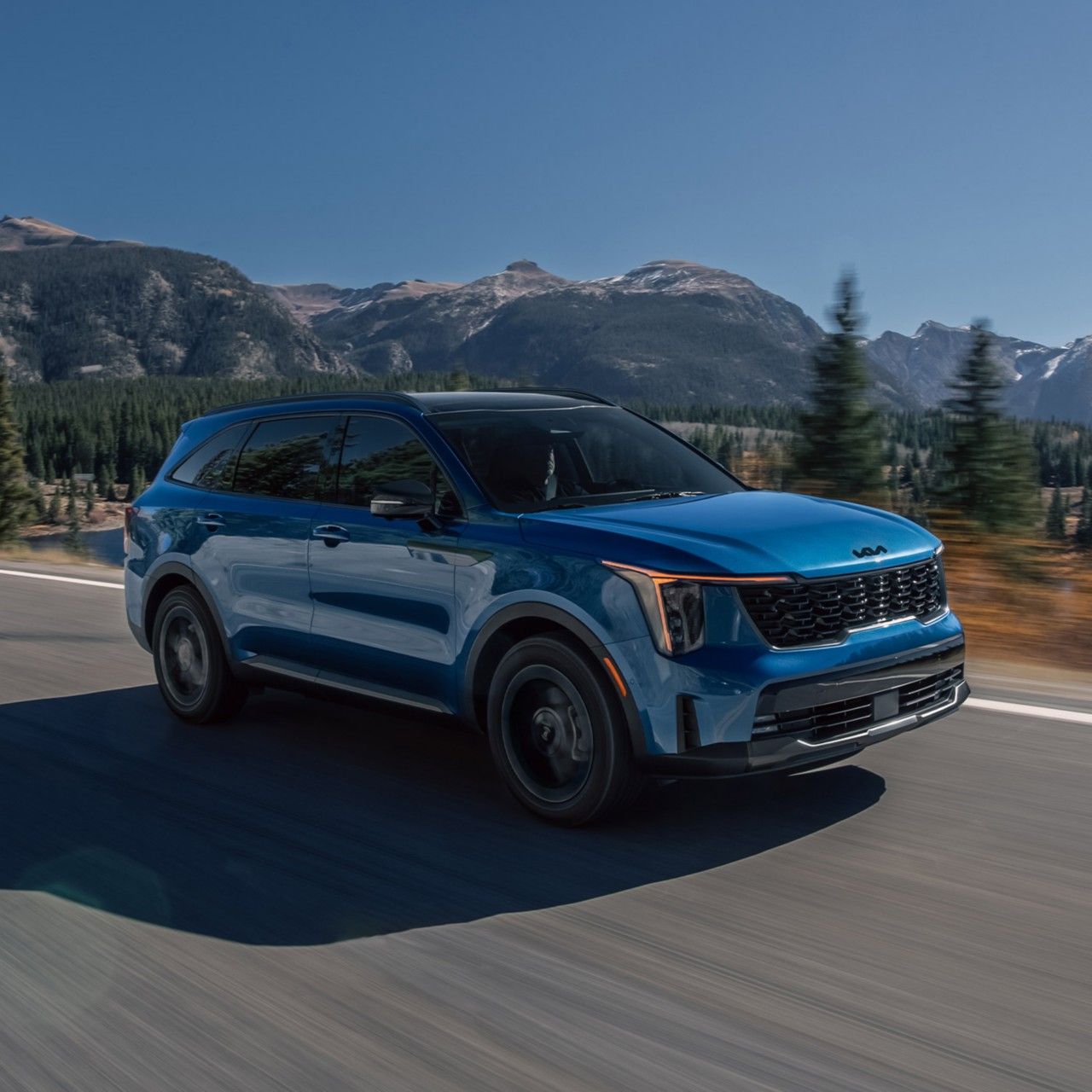 Scenic photograph of the 2026 Kia Sorento Hybrid SUV in blue driving along a road with pine trees and mountains in the back, highlighting the LED headlights and all-wheel drive.