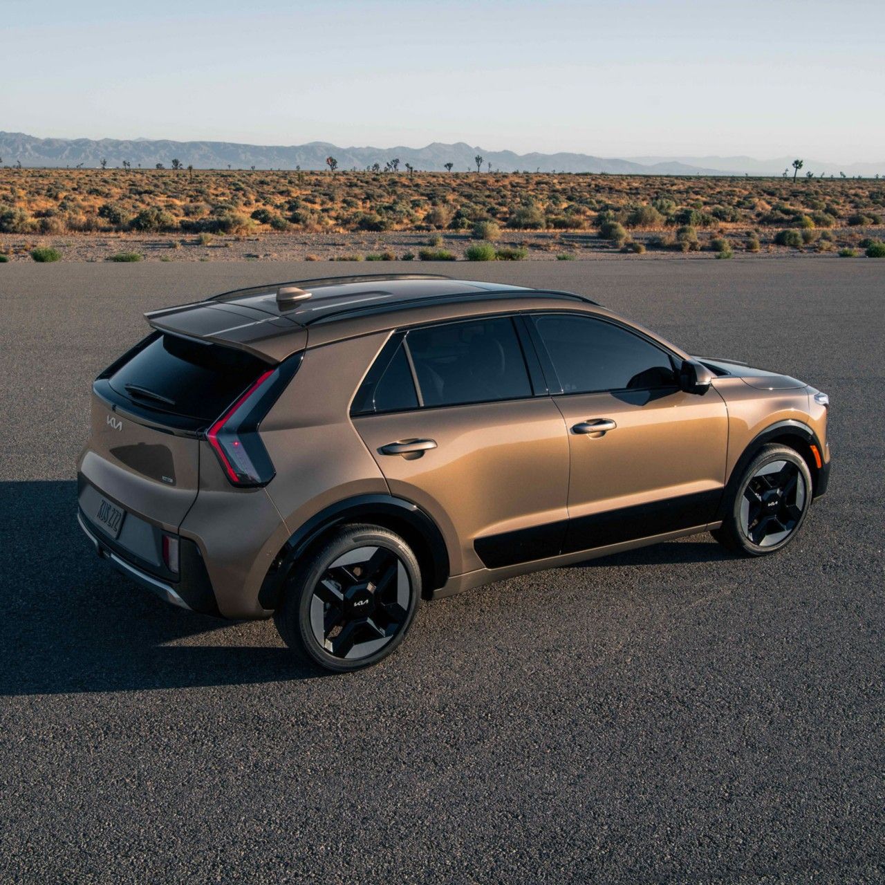 Side-angle photo of the 2027 Kia Niro Hybrid in brown, showcasing its taillights, stylish exterior, and tinted windows. The SUV is parked on the street with desert views and mountains behind it.