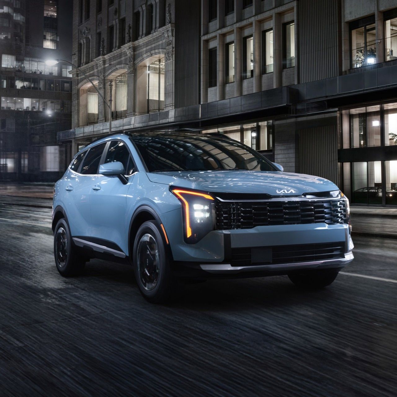 Front three-quarter view of the 2027 Kia Sportage Hybrid in blue driving on an urban street at night with buildings in the background, highlighting the compact SUV’s vertical LED headlights.