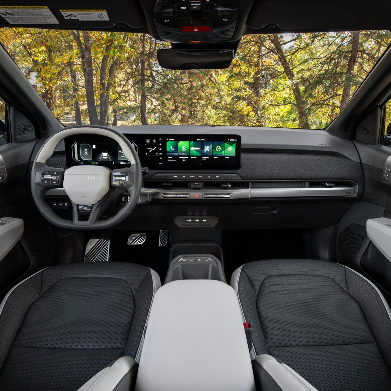 Interior view of the 2027 Kia EV3 compact electric SUV's steering wheel, dashboard, digital display panel, center console, and front seats, showcasing the modern features and styling inside the EV. A lush forest is visible through the windshield.