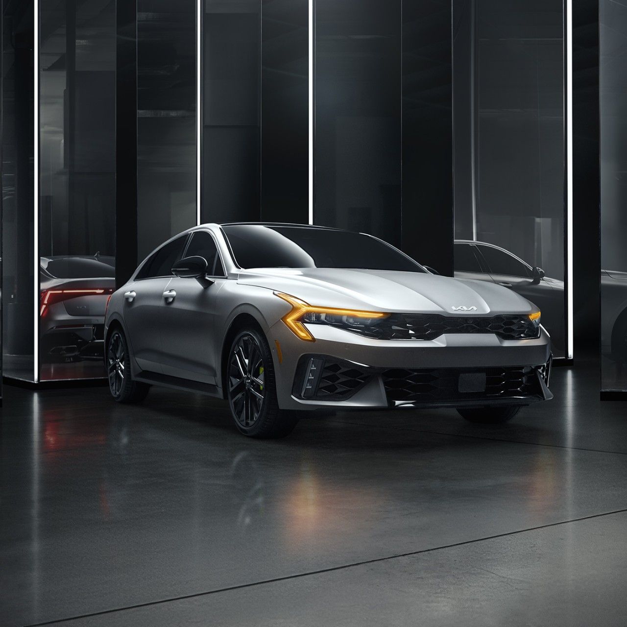 A gray 2027 Kia K5 parked in an all-white room, featuring the sedan’s LED headlights, tinted windows, bold front grille, and gloss black wheels.
