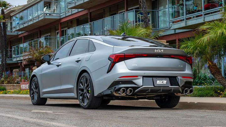 Rear three-quarter view of the 2027 Kia K5 in gray parked in an urban setting, showcasing its taillights, sporty rear design, and four exhaust tips.