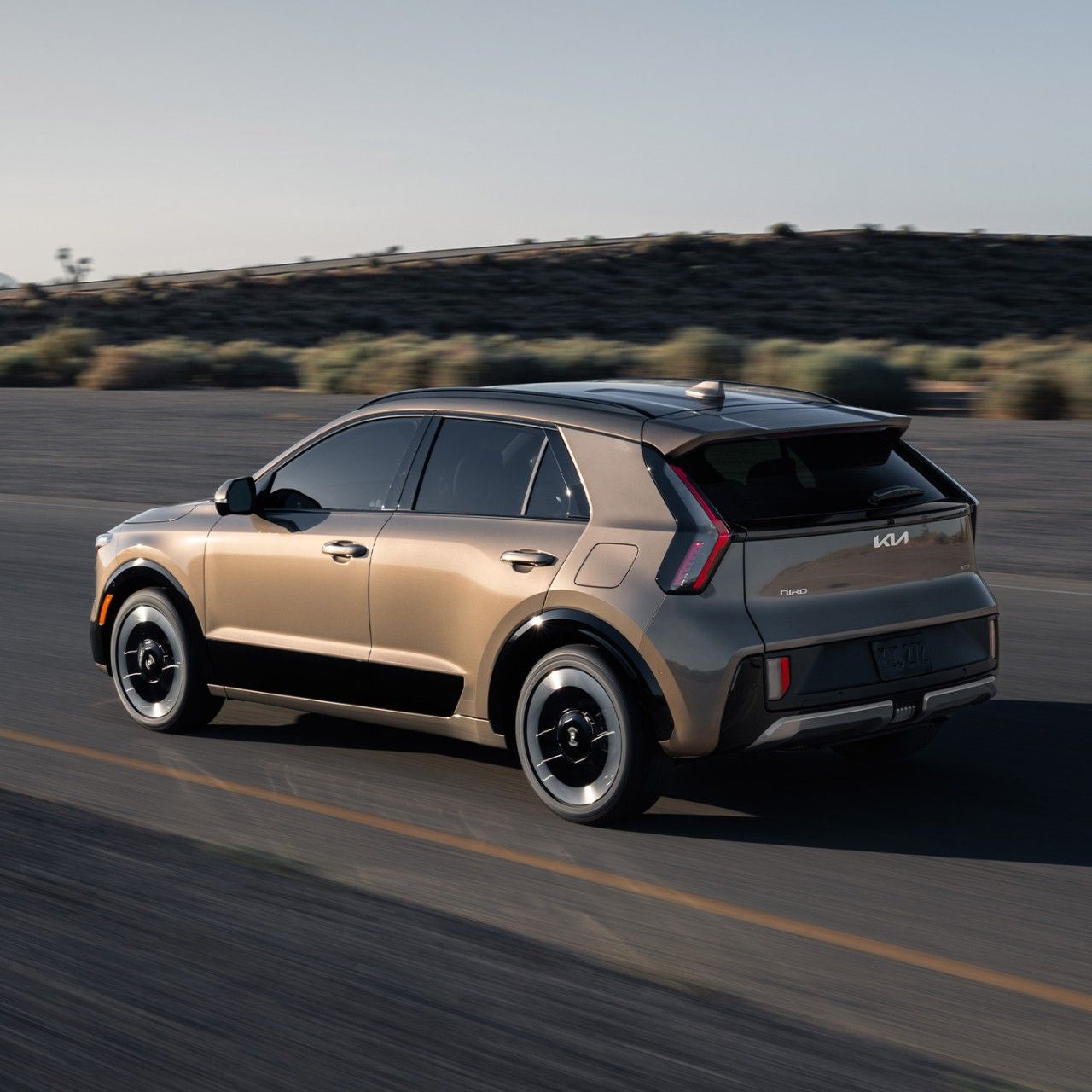Rear angle photograph of the 2027 Kia Niro Hybrid Electric SUV driving over an outdoor road with desert vegetation in the background.
