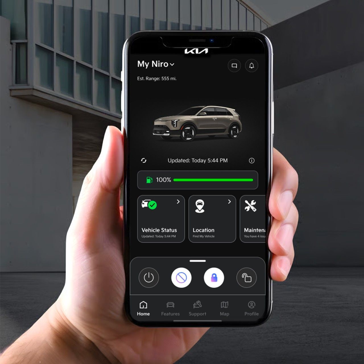 A hand holding a cellular phone, which has the Kia Connect app onscreen. The app is displaying a Kia vehicle and various status and location information. Building architecture is visible in the background. 