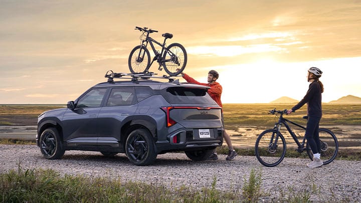 A dark gray 2027 Kia Seltos is parked on a gravel path at sunset, with two people loading bicycles onto the roof rack. One person stands beside the SUV holding a bike, while the other secures a second bike on top, with open terrain and distant hills in the background.